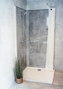 Moderne Eckdusche mit Glasabtrennung, grauen Wandfliesen, Handbrause und dekorativer Grünpflanze im Korb.