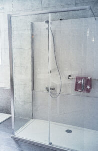 Moderne Dusche mit Glas-Schiebetür, Handbrause, weiß gefliester Wand und Handlauf mit Handtuch.