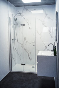Moderne Dusche mit Glasabtrennung, Marmoroptik-Wänden, Regendusche und angrenzendem weißen Waschtisch mit Chromarmatur.