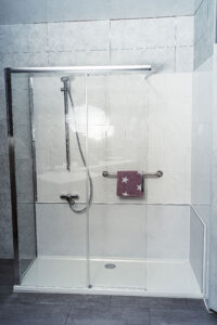 Barrierearme Dusche mit klarer Glasfront, Schiebetür, Handbrause und Haltegriff mit Handtuchhalter.