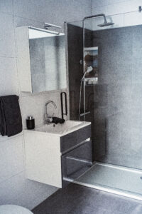 Modernes Badezimmer mit grauer Glasdusche, Regenduschkopf, weißem Waschtisch mit Spiegel und schwarzen Accessoires.