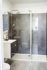 Badezimmer mit großer Dusche, Glasabtrennung und grauen Wandfliesen, ergänzt durch ein weißes Waschbecken mit Unterschrank.