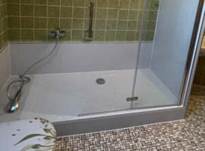 Barrierearme Dusche mit niedriger Einstiegskante, grüner Fliesenwand, Handbrause und transparenter Glastür, auf Mosaikfliesenboden.