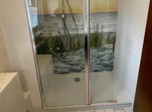 Barrierearme Dusche mit Glas-Schiebetüren, integrierter Sitzfläche und großformatigem Wandmotiv eines Strandes mit Dünen und Meer im Sonnenuntergang.