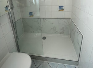 Barrierearme Dusche mit Glasabtrennung, weißer Duschwanne und Wandverkleidung in eleganter Marmoroptik, kombiniert mit weißen Fliesen und kleinen maritimen Dekorfliesen.