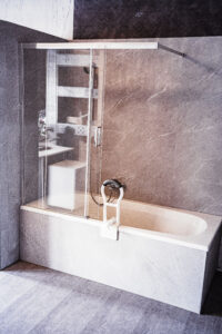 Badewanne mit seitlicher Glasabtrennung, Handbrause und montierter Einstiegshilfe, um den Zugang zur Wanne zu erleichtern, vor grauer Wandverkleidung in Marmoroptik.
