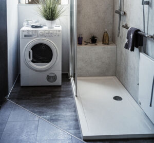 Barrierearme Dusche mit Glasabtrennung, integrierter Sitzbank, Handbrause, Haltegriff und angrenzendem Platz für eine Waschmaschine.