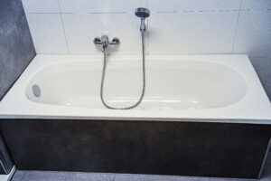 Weiße Badewanne mit Wandarmatur und Handbrause, vor einer Wand mit weißen und grauen Fliesen, Wannenverkleidung in dunkler Steinoptik.