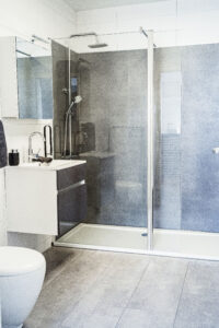 Modernes Badezimmer mit bodengleicher Glasdusche, grauen Wand- und Bodenfliesen, weißem Waschtisch und Wandspiegel.