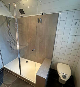 Moderne, barrierearme Dusche mit großer Glasabtrennung, integrierter Sitzfläche, Handbrause und hellen Wandverkleidungen in Holzoptik, neben einem wandhängenden WC.