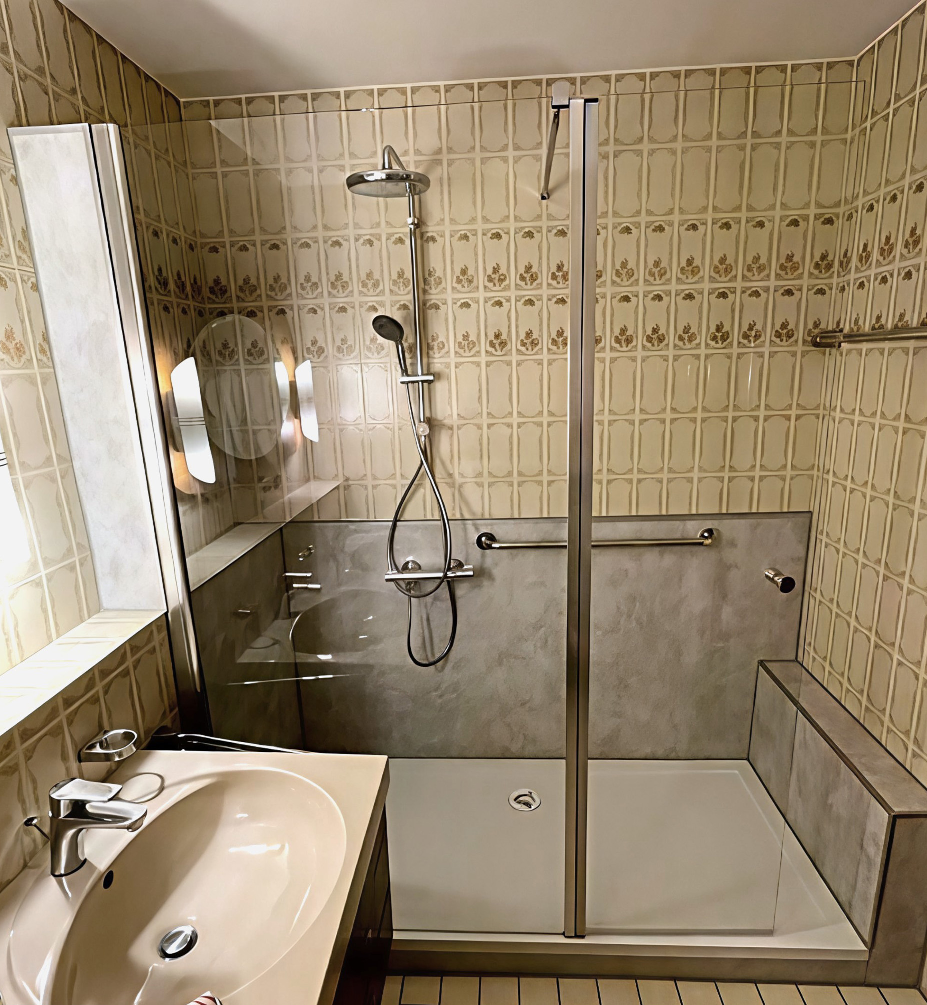 Barrierearme Dusche mit Glasabtrennung, grauer Wandverkleidung, Regenduschkopf, Handbrause, Haltegriffen und integrierter Sitzbank, neben einem Waschtisch.
