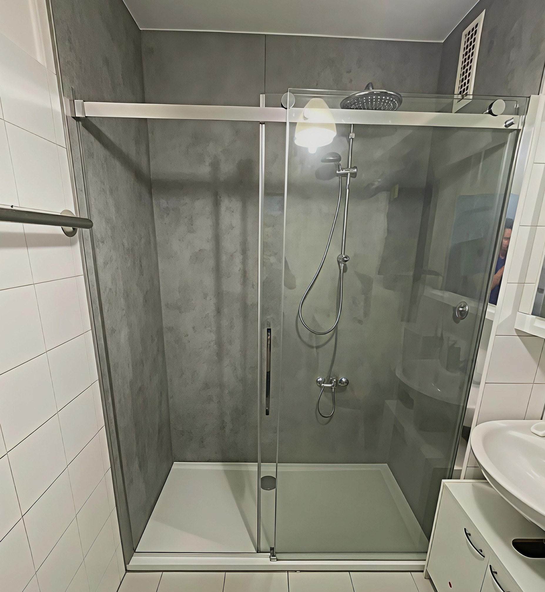 Moderne, barrierearme Dusche mit grauer Wandverkleidung in Betonoptik, großer Glas-Schiebetür, Handbrause und Regenduschkopf, neben einem weißen Waschtisch.