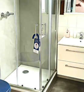 Barrierearme Dusche mit Eckzugang, Glasabtrennung und verchromten Griffen, neben einem modernen weißen Waschschrank mit Spiegelschrank.