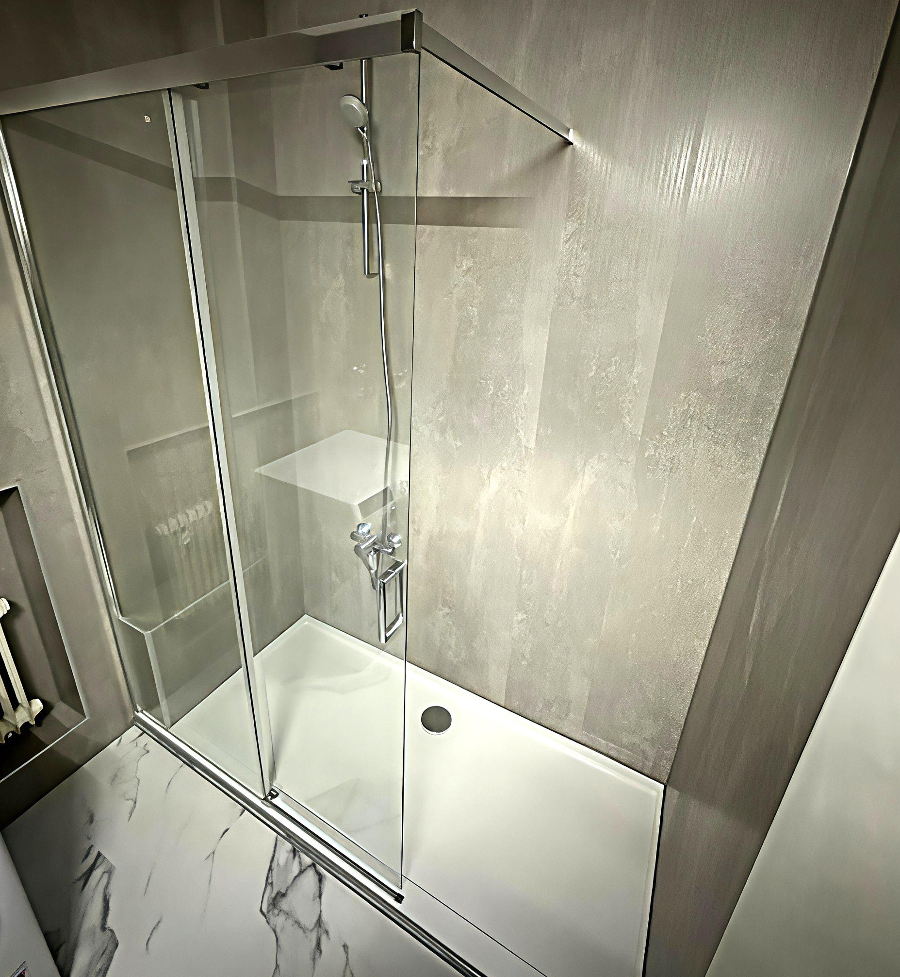Moderne, barrierearme Dusche mit Glas-Schiebetür, grauer Wandverkleidung in Betonoptik, weißer Duschwanne und integrierter Ablagefläche.