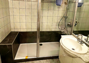 Barrierearme Dusche mit Glasabtrennung, integrierter Sitzbank und dunkler Marmoroptik-Wandverkleidung, neben einem weißen Waschtisch mit Marmorplatte.