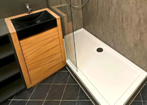 Barrierearme Dusche mit Glasabtrennung, heller Duschwanne und grauer Wandverkleidung, neben einem modernen Waschtisch mit Holzfront und schwarzem Becken.