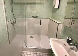 Barrierearme Dusche mit Glasabtrennung, integrierter Sitzbank, Handbrause und Haltegriff, neben einem weißen Waschbecken und Spiegelschrank.