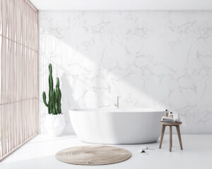 Helles, minimalistisches Badezimmer mit freistehender weißer Badewanne, Marmorwand, Kaktus und kleinem Holzbeistelltisch.