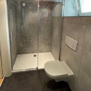 Eckdusche mit Glasabtrennung, grauer Wandverkleidung und wandhängendem WC – barrierearm umgebaut.