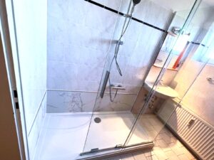 Barrierearme Dusche mit Glas-Schiebetür, weißer Duschwanne und Wandverkleidung in eleganter Marmoroptik.