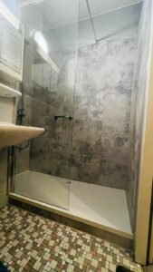 Barrierearme Dusche mit Glasabtrennung, grauen Wandpaneelen in Betonoptik und kontrastreichem Mosaikfliesenboden.