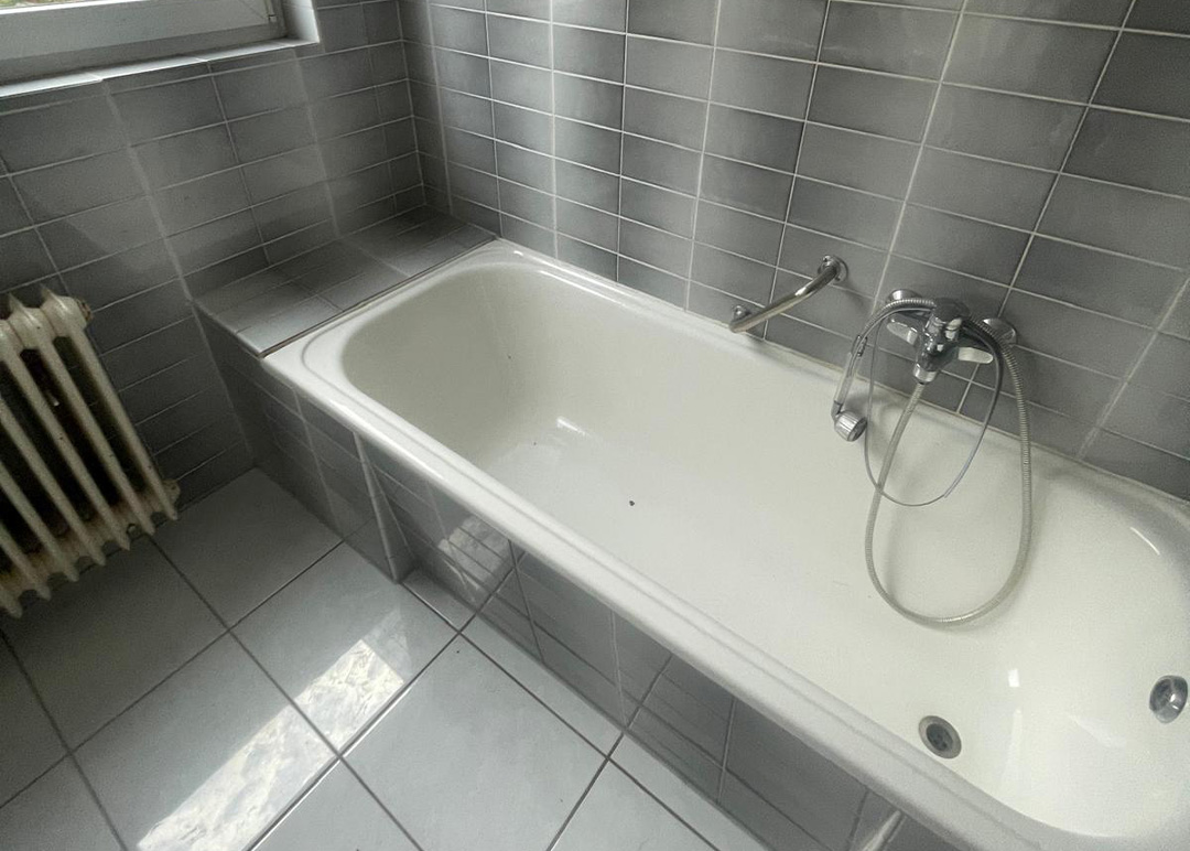 Bad mit klassischer Badewanne, grauen Fliesen und hohem Einstieg vor dem Umbau.