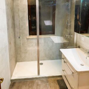 Barrierearme Dusche mit Glas, grauer Wandverkleidung und Fensterbank, neben weißem Waschtisch.