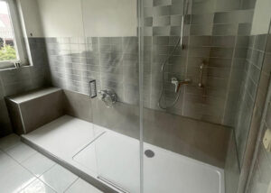 Barrierearme Walk-In-Dusche mit Glasabtrennung, Sitzbank links, Haltegriff und grauen Wandfliesen.